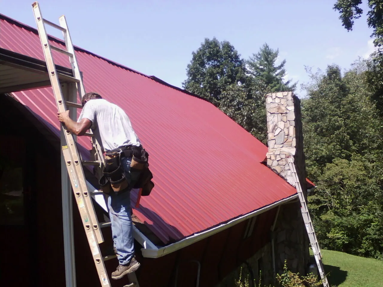 Roofer on a ladder beside a red metal roof and stone chimney — Soffit & Fascia Repair by Roofing Brothers in Licking